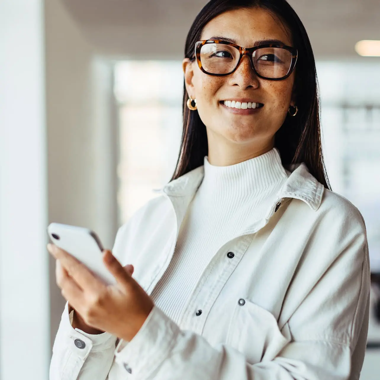 A woman with glasses is smiling while holding a phone. She is indoors, wearing a shirt with sleeves and has a collar. The background features a wall.