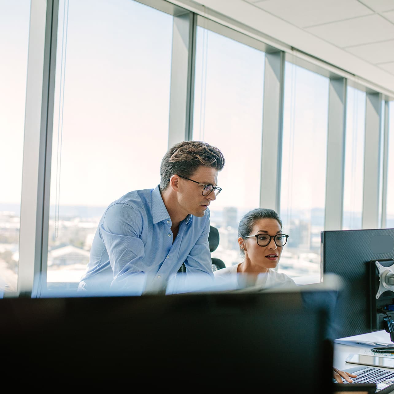 A man and woman are sitting in an office building, looking intently at a computer screen. The setting includes a window with natural light coming in, and they are both dressed in professional clothing. The man is wearing glasses and they appear engaged in their task, surrounded by office decor.